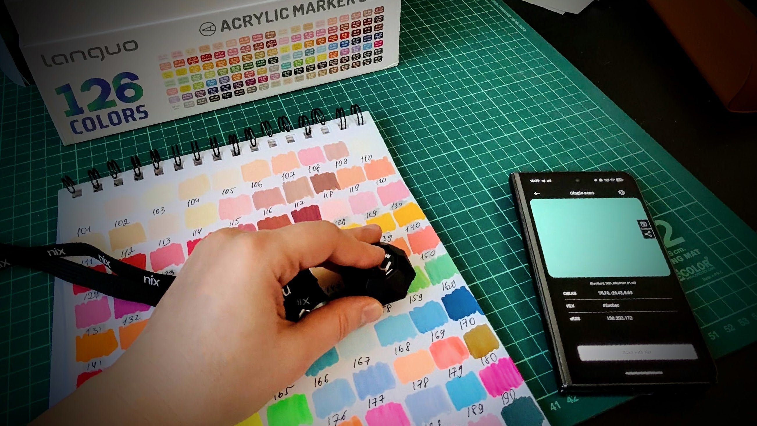 Color swatch book with a hand holding a marker, phone displaying color samples, and marker box in the background. Nix sensor measures color for pen plotter art library of tools.