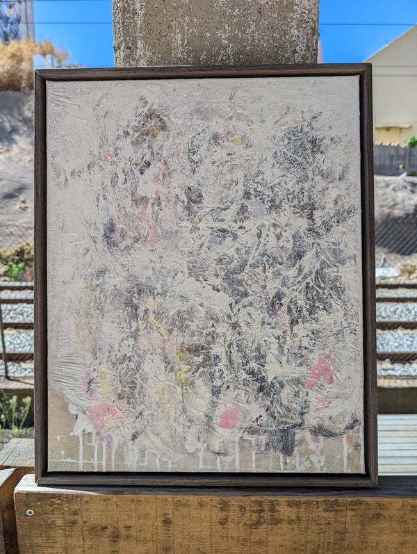an abstract painting with a mix of white, gray, and pink colors, displayed in a wooden frame on a wooden surface.