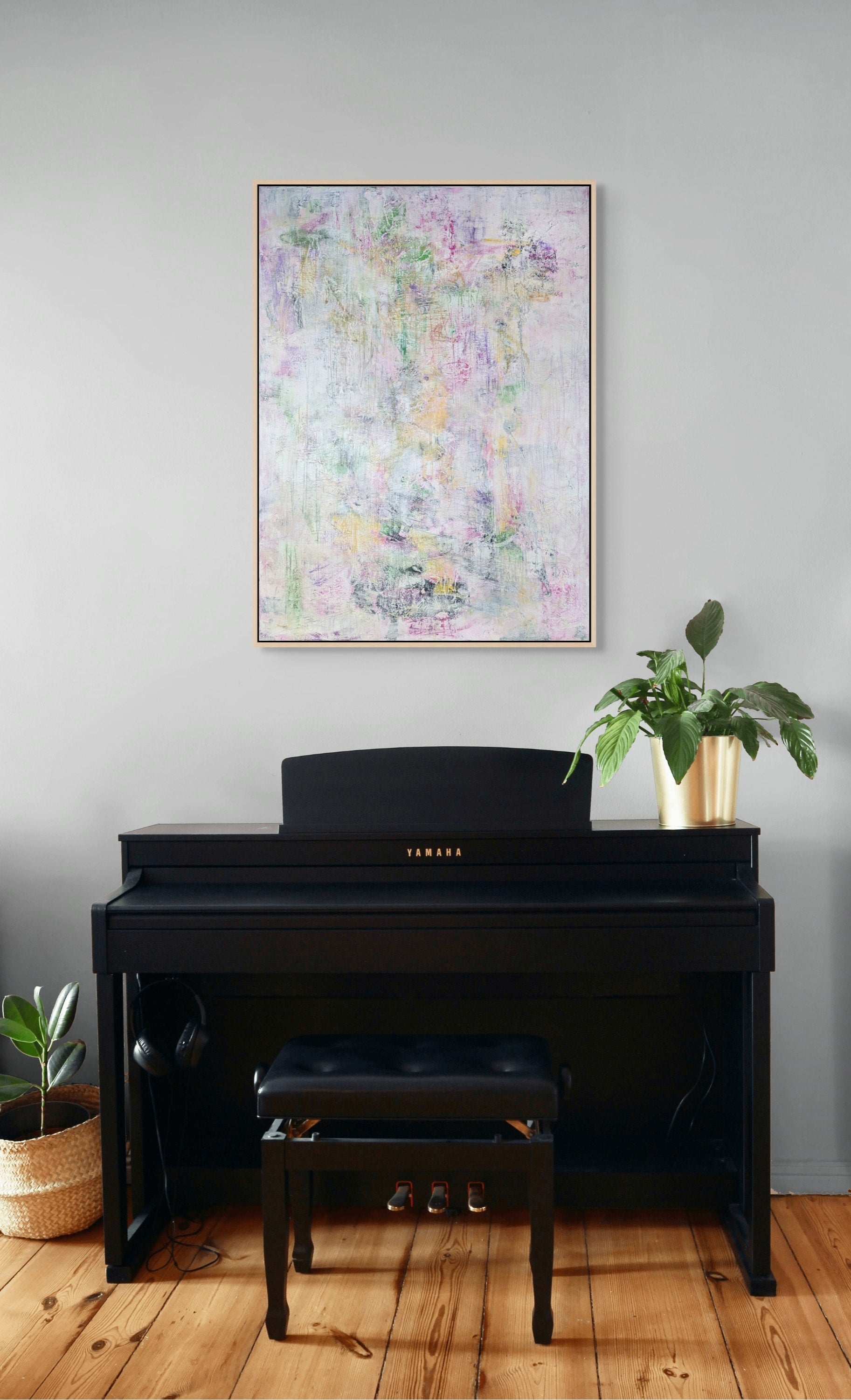 a Yamaha piano with a black frame, a black stool, and a potted plant on the floor next to the piano. The piano is positioned in front of a gray wall, and a framed abstract painting is hanging above it.